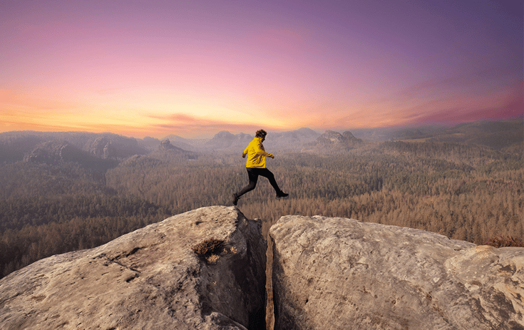 Woman jumping over a rock crevice with a pink sunset